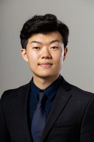 Professional headshot of a young Asian man (Daniel Jung) in a dark suit with a blue tie and shirt, smiling confidently at the camera with a light grey background.